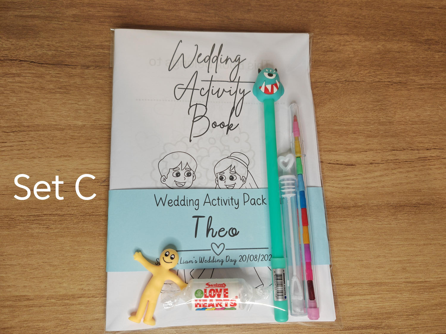 A set of children's wedding activity packs, including a coloring book, a changeable crayon, a novelty pen, and a small toy, displayed on a wooden table.