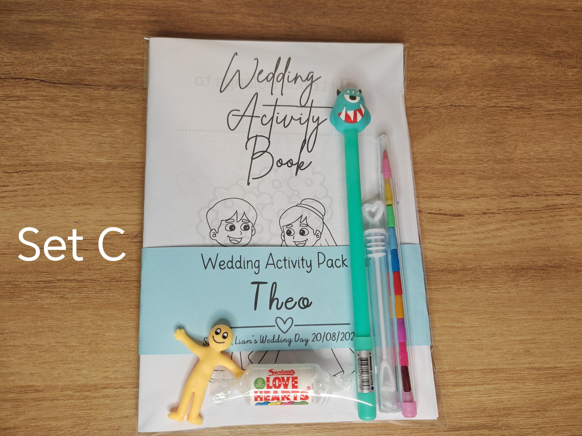 A set of children's wedding activity packs, including a coloring book, a changeable crayon, a novelty pen, and a small toy, displayed on a wooden table.